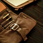 A partially unrolled tool roll with a brown waxed canvas exterior, aluminum hook fastener, and multiple pockets containing pens and other tools.