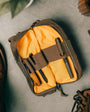 A brown and yellow venture pouch with elastic loops and pockets, alongside a pair of shoes and a bag, suggesting it can be carried in a bag or used as a crossbody bag.