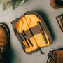 A brown and yellow venture pouch with elastic loops and pockets, alongside a pair of shoes and a bag, suggesting it can be carried in a bag or used as a crossbody bag.