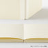 A cream-colored lined notebook shown open, with a folded view of the same notebook above it on a clean white background. Small Japanese text appears in the bottom right corner.