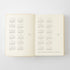 An open cream-colored planner or notebook showing printed calendar pages with dates and Japanese text, photographed against a light gray background.