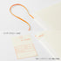 An off-white hardcover notebook or journal shown on a white background, with an orange loop tab, a small orange-red 'DIARY' label, and an index sticker sheet with Japanese text. The notebook cover has a subtle woven texture.