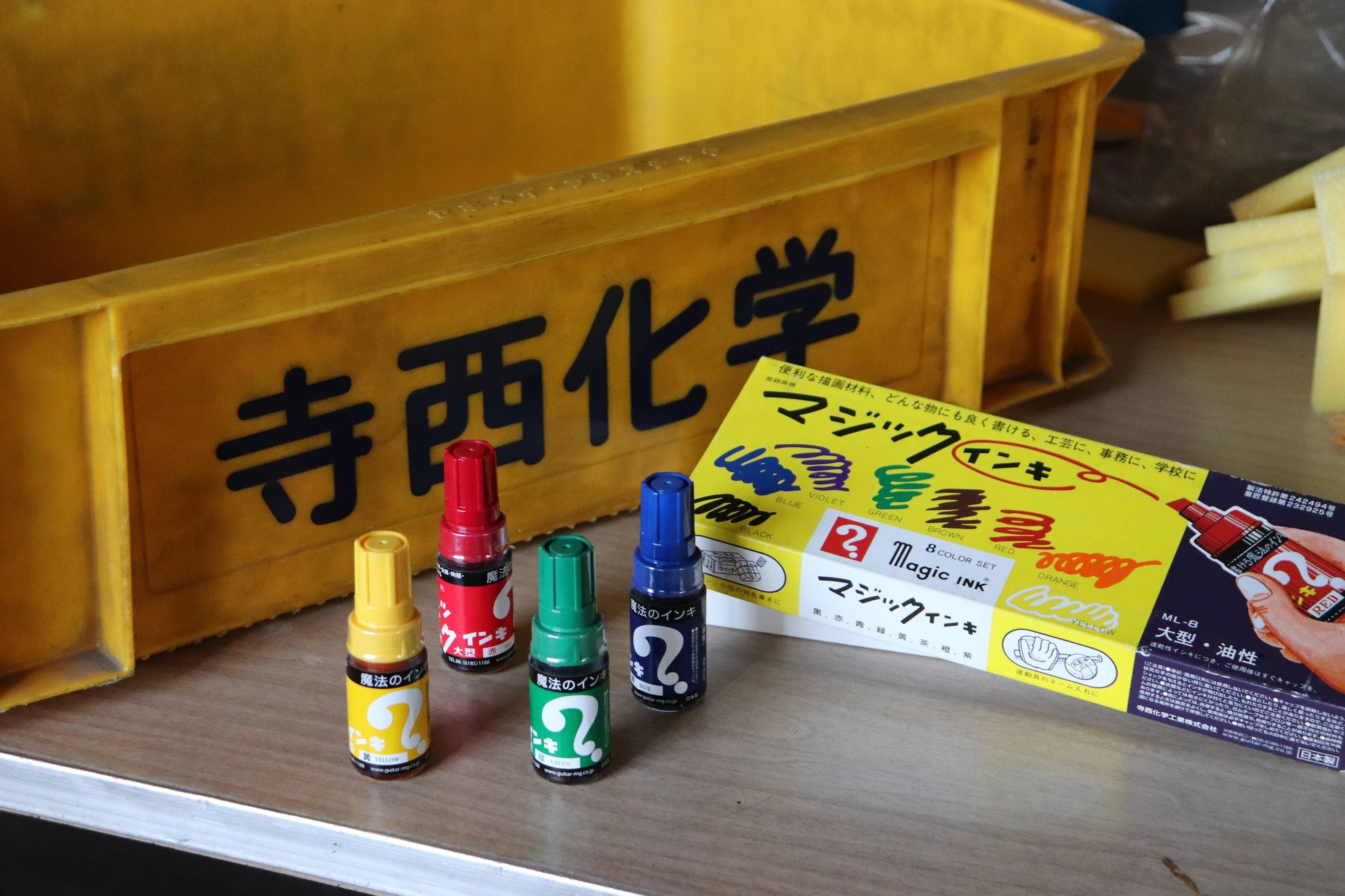 Four small bottles with colorful caps and a box on a wooden surface with a yellow sign in the background.