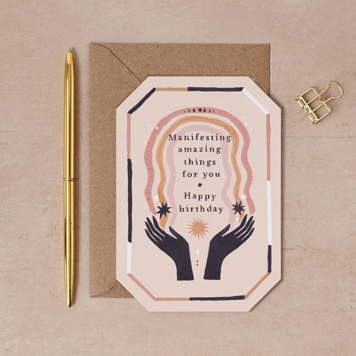Greeting card with hands holding a heart design and birthday message, placed on a beige surface with a pen and gold clip.