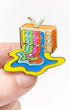 A small enamel pin shaped like a retro television with colorful dripping liquid patterns, held between fingers against a plain white background.