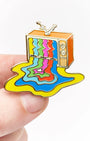 A small enamel pin shaped like a retro television with colorful dripping liquid patterns, held between fingers against a plain white background.