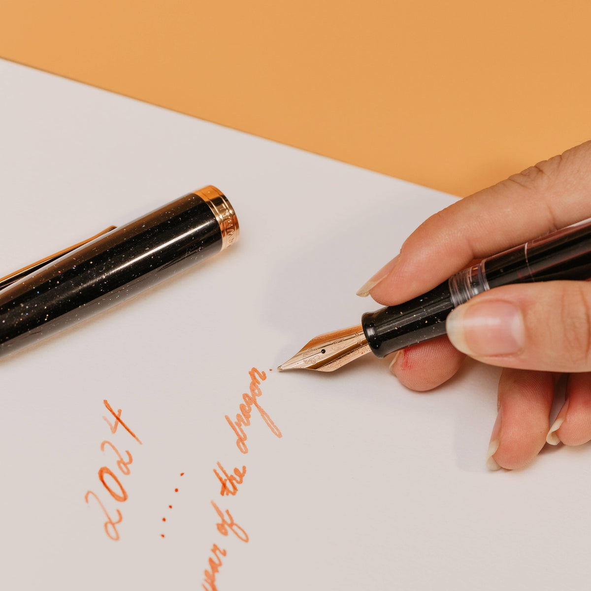Hand holding a fountain pen writing on paper with an orange background