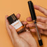 A hand holds a small bottle of Honey Bomb Nectar 2 Syphon ink next to a black and clear fountain pen with gold trim, against a solid orange background.