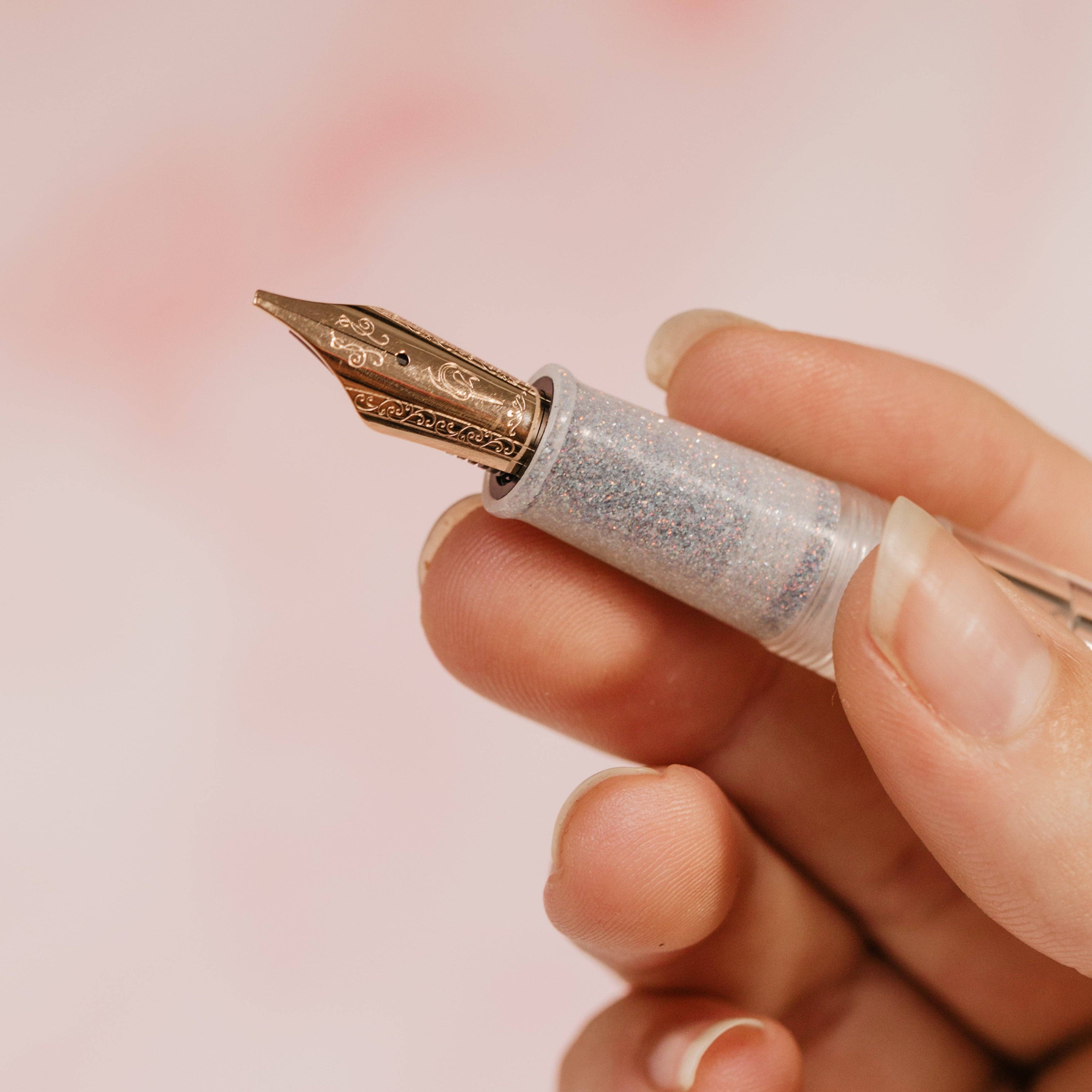 Fountain pen with decorative nib held in a hand against a soft pink background