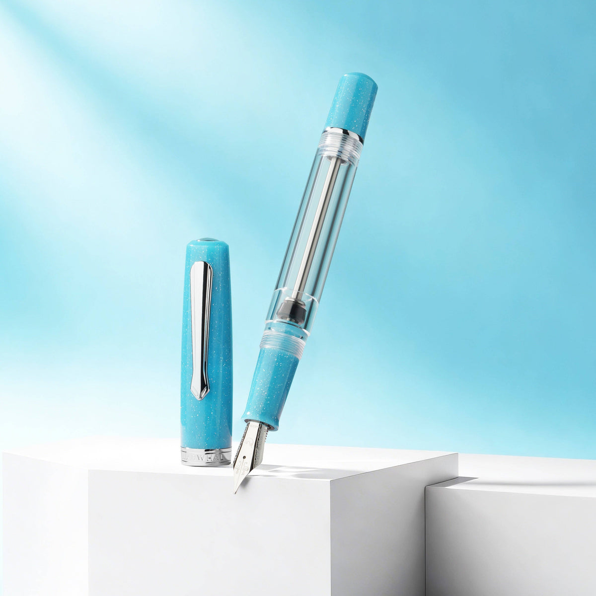 A light blue fountain pen with a transparent barrel and silver clip, shown with its cap removed and standing on white display blocks against a soft blue background.