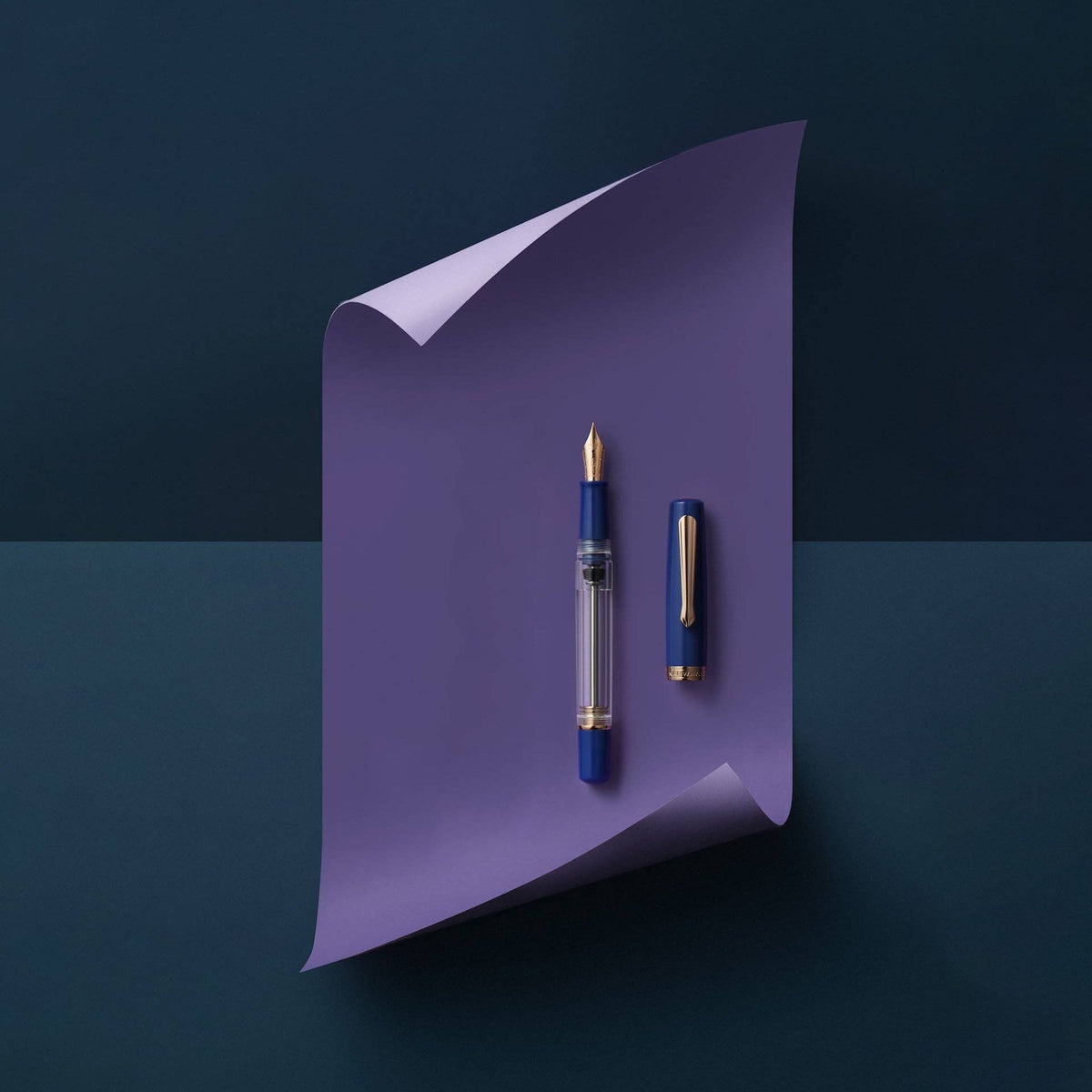 A blue and gold fountain pen with its cap removed, displayed on a curled purple sheet of paper against a dark blue background.