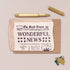 A styled flat-lay featuring a brown kraft envelope with a cream-colored paper newspaper print that reads 'WONDERFUL NEWS,' a gold pen, a gold cap, and a few postage stamps on a light beige background.
