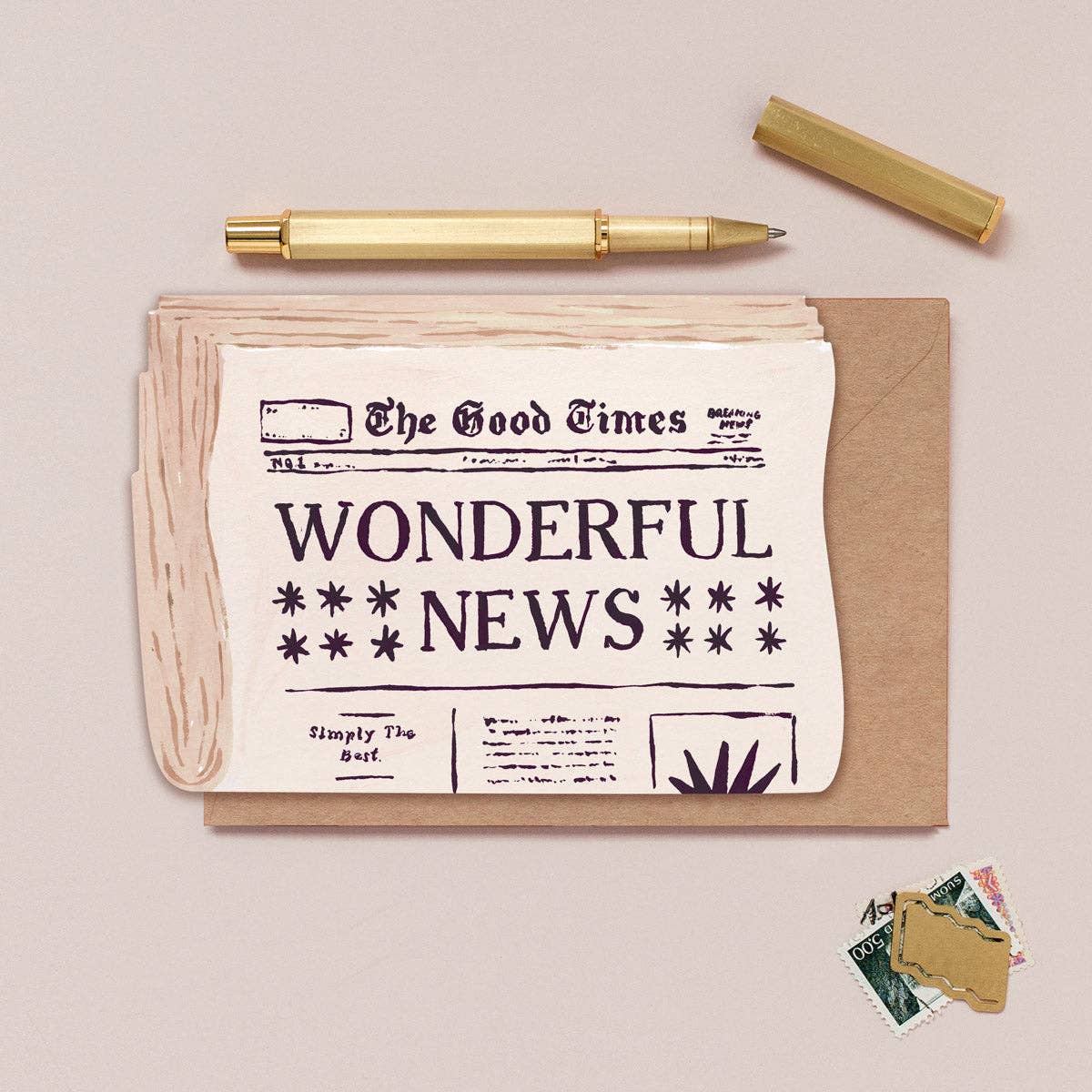 A styled flat-lay featuring a brown kraft envelope with a cream-colored paper newspaper print that reads 'WONDERFUL NEWS,' a gold pen, a gold cap, and a few postage stamps on a light beige background.