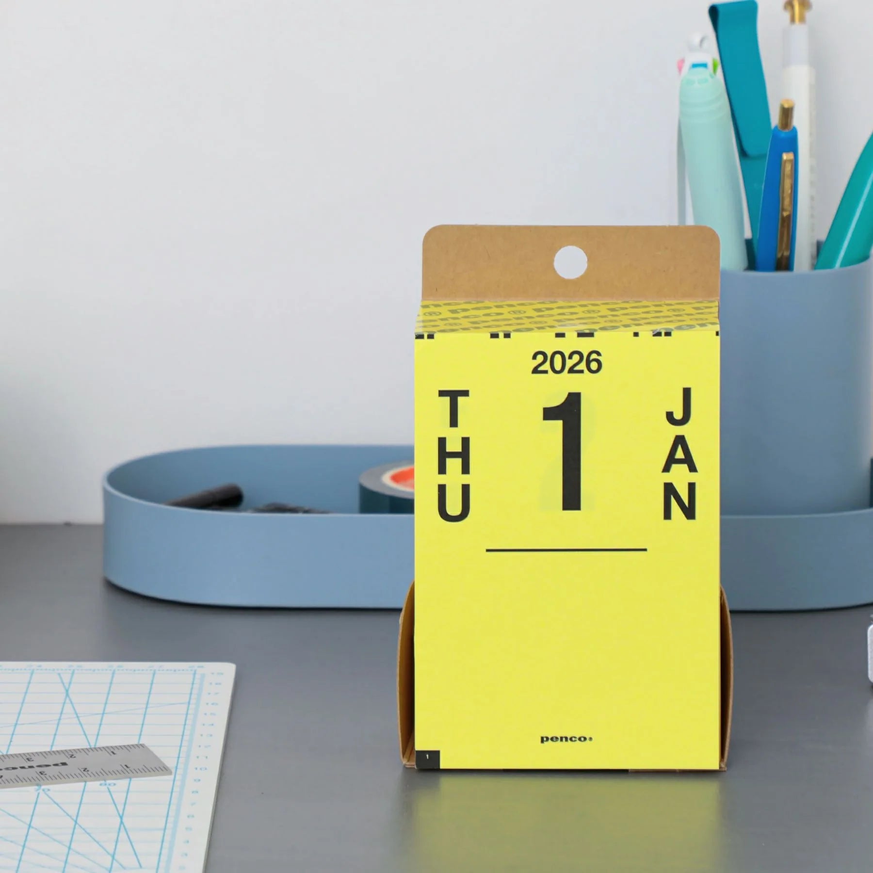 Yellow desk calendar for 2026 on a gray surface with office supplies in the background.