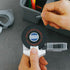 A close-up of a person using a Pencq brand black rubber tape measure with a white plastic handle and clear plastic casing, holding a black tape label that reads 'SUGA'.