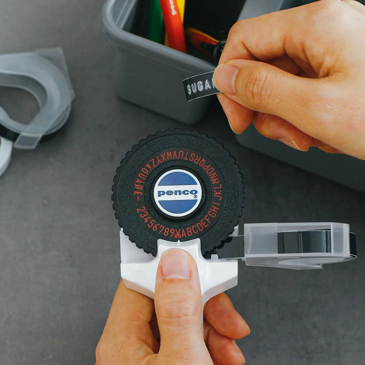 A close-up of a person using a Pencq brand black rubber tape measure with a white plastic handle and clear plastic casing, holding a black tape label that reads 'SUGA'.