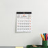 A 2027 January wall calendar hanging on a light gray wall above a desk with a green cup of colored pencils.