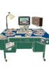 A vintage-style desk or control console with a blue top, green base, multiple circular dials, a small screen, a lamp, and a speaker, isolated on a white background.