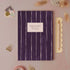 A weekly planner notebook with a dark purple cover featuring vertical white stripes and a white label in the center that reads 'WEEKLY PLANNER' and 'Made in England'. The notebook is styled in a flat-lay arrangement on a dusty pink background with stationery props including a wooden ruler, gold binder clips, washi tape, and dried grass.
