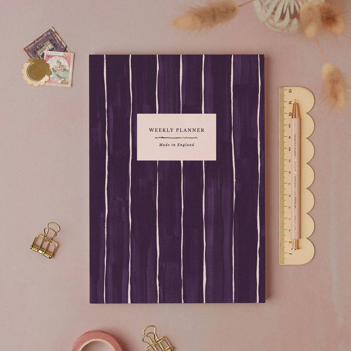 A weekly planner notebook with a dark purple cover featuring vertical white stripes and a white label in the center that reads 'WEEKLY PLANNER' and 'Made in England'. The notebook is styled in a flat-lay arrangement on a dusty pink background with stationery props including a wooden ruler, gold binder clips, washi tape, and dried grass.