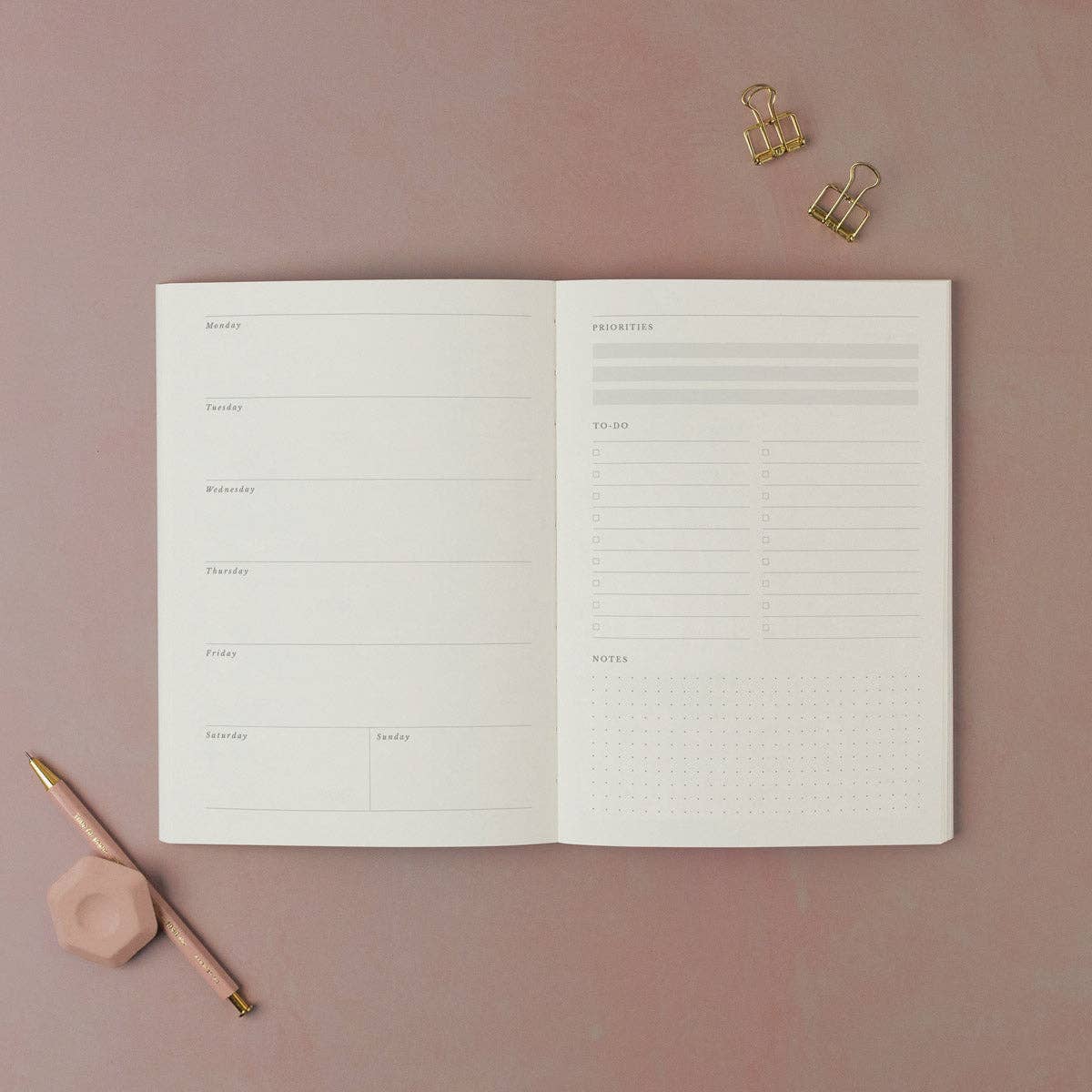 Open notebook with lined pages on a pink background, accompanied by a pen and clips.