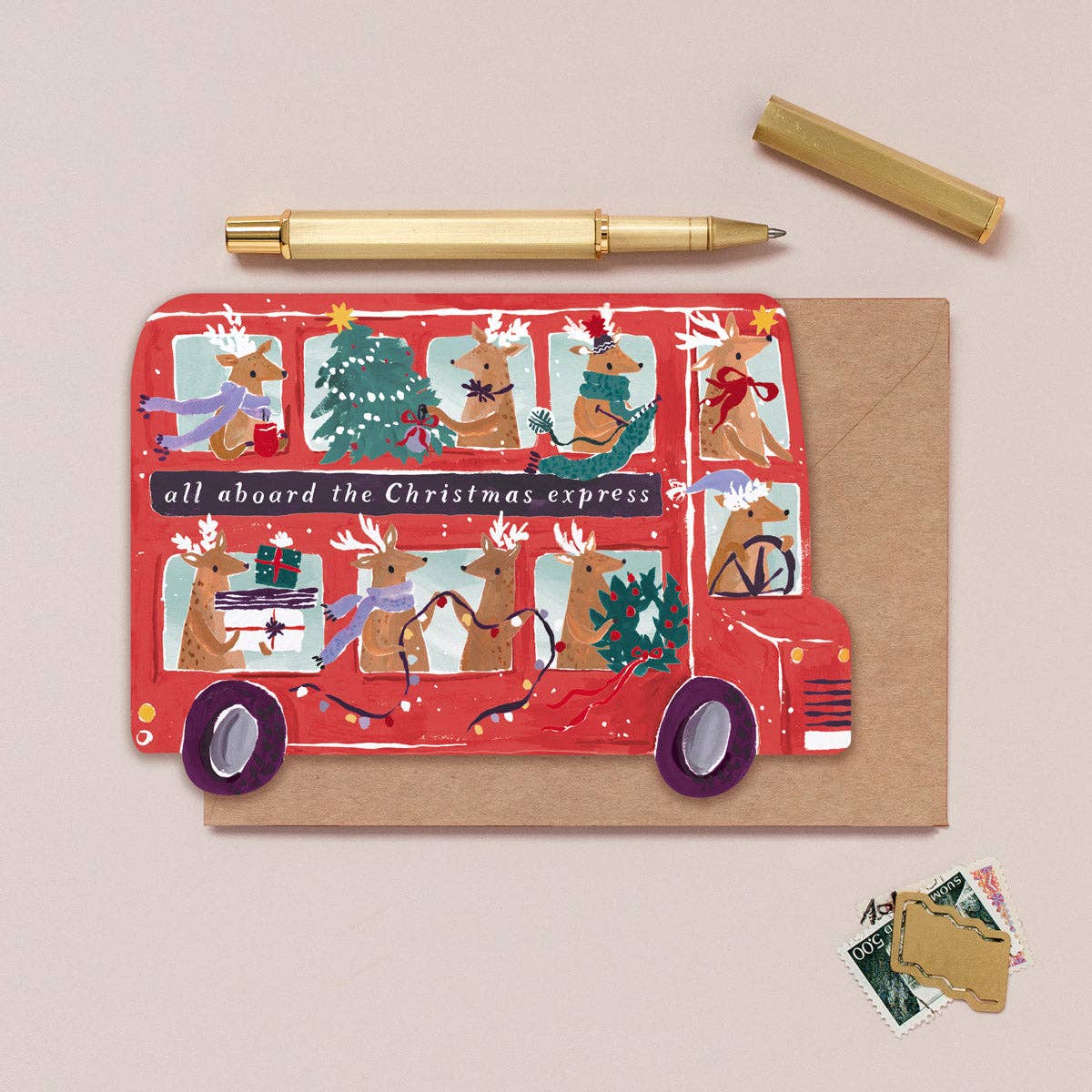 Christmas-themed card with red bus design and reindeer, accompanied by a pen and small envelopes on a beige background.