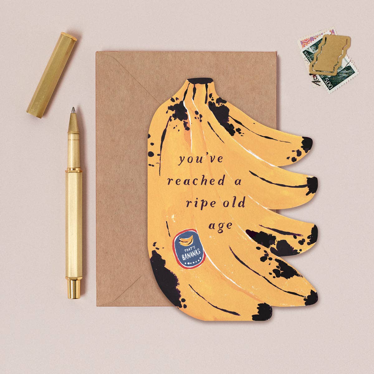 Greeting card with banana design and text, accompanied by a pen and envelope on a beige background.