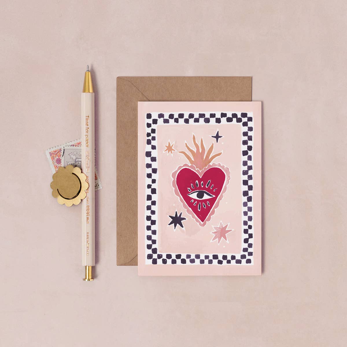 A flat-lay product photo of a 'Holy Heart' greeting card, shown with its brown kraft envelope and a gold pen on a textured beige background.