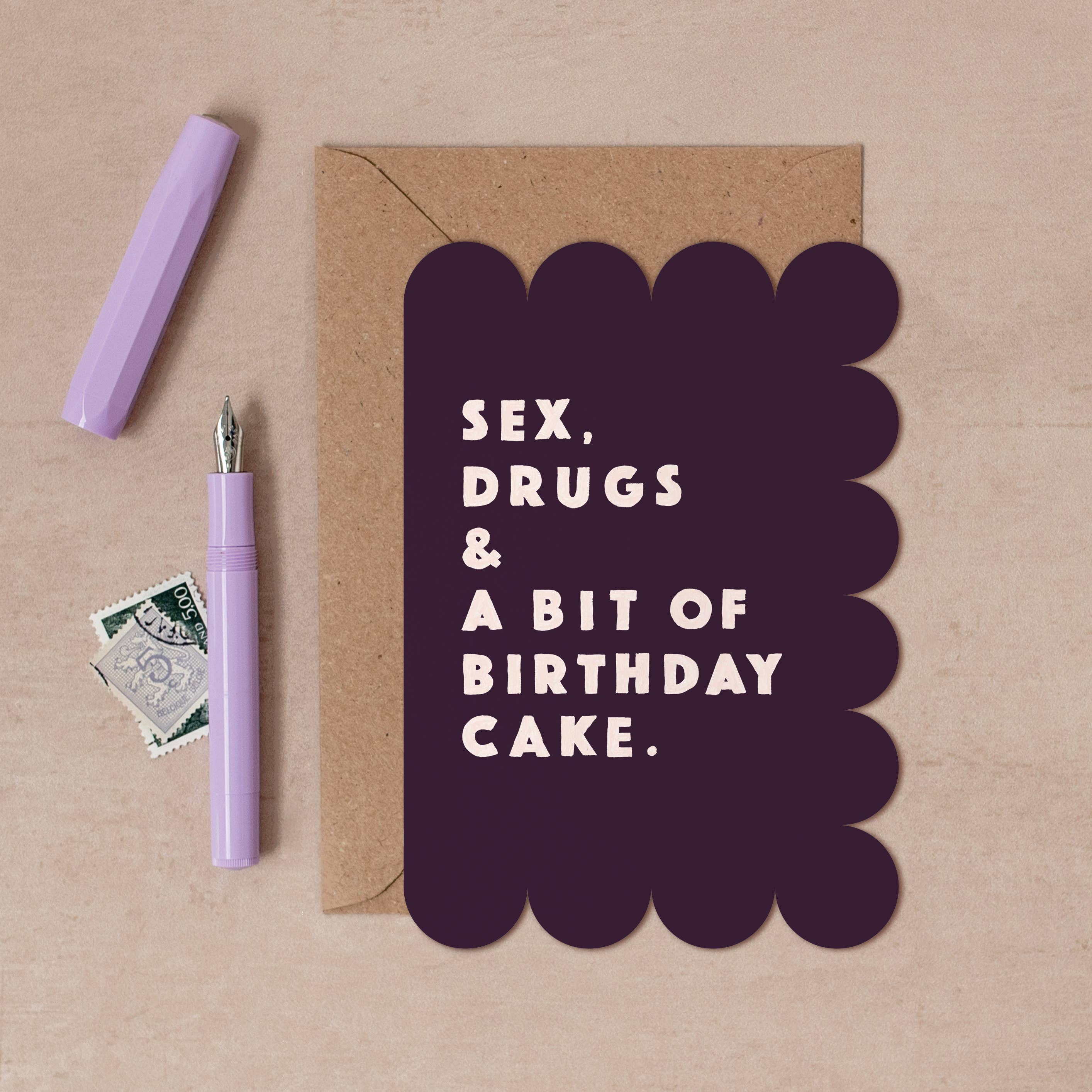A flat-lay of a novelty birthday card with a dark purple scalloped border and the text 'SEX, DRUGS & A BIT OF BIRTHDAY CAKE.' in white, shown with a kraft envelope, a lavender pen, and a small postage stamp on a beige background.