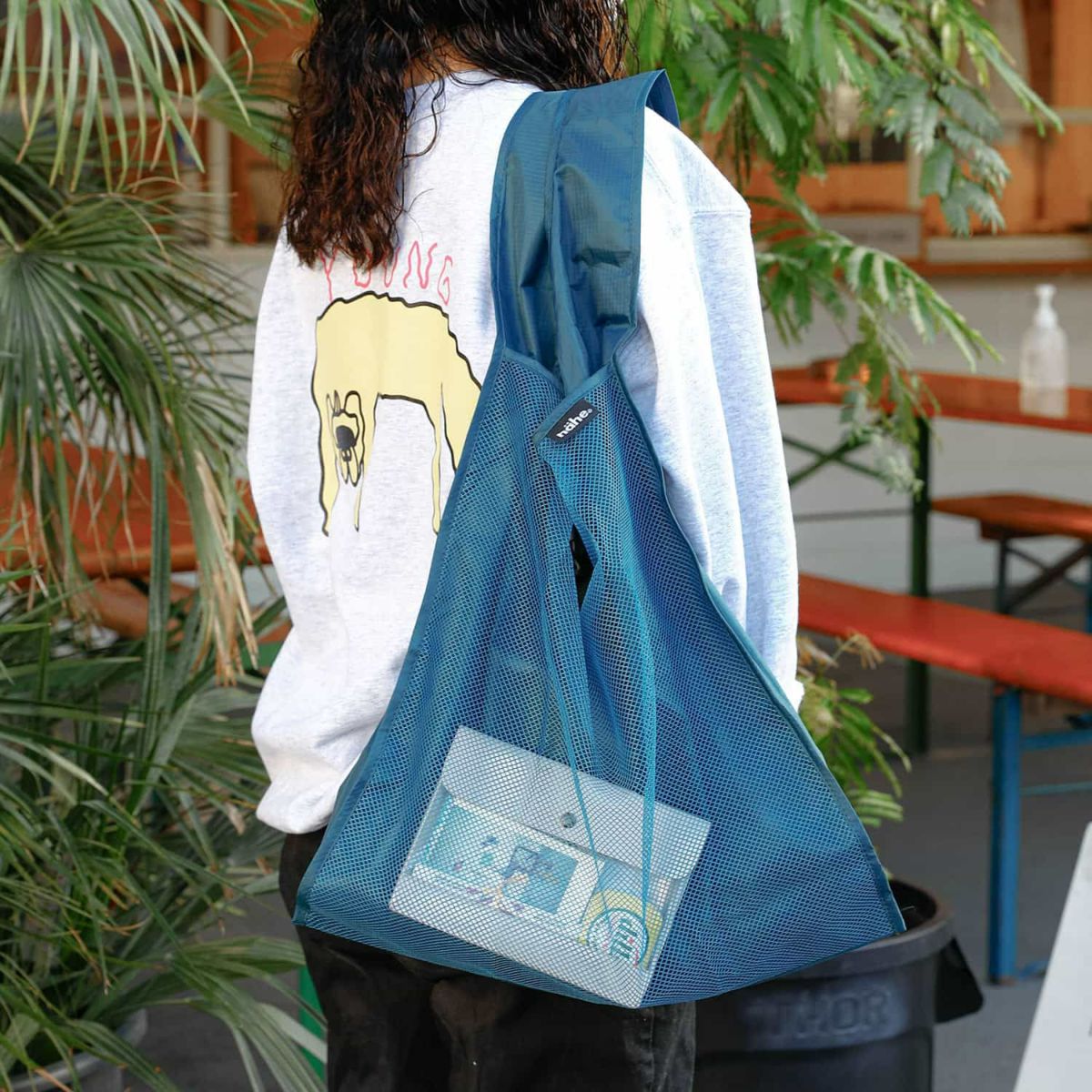 Person wearing a blue mesh bag over their shoulder in an outdoor setting with plants and furniture.