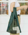 Person holding a green mesh bag with a white shirt and beige pants in front of a window.