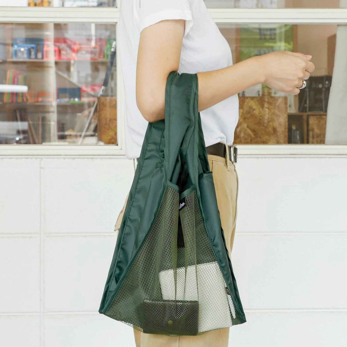 Person holding a green mesh bag with a white shirt and beige pants in front of a window.