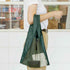 Person holding a green mesh bag with a white shirt and beige pants in front of a window.
