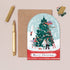Christmas card with illustration of people decorating a tree, next to a pen and envelope on a beige background