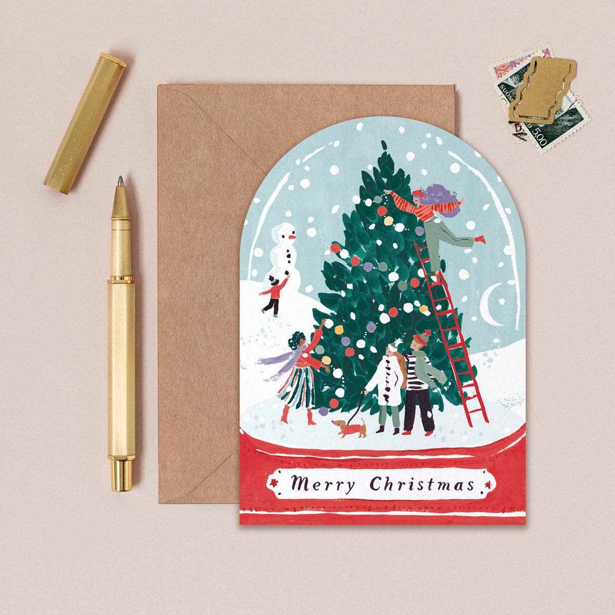 Christmas card with illustration of people decorating a tree, next to a pen and envelope on a beige background