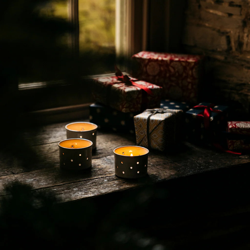 Three lit candles in decorative holders on a wooden surface with wrapped gifts in the background.