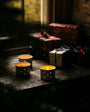 Three lit candles in decorative holders on a wooden surface with wrapped gifts in the background.
