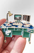 A miniature desk-themed enamel pin held in a person's hand against a light gray background. The pin features a blue desk with green drawers, gold-tone metal outlines, a silver desk lamp, a computer monitor, and a speaker.