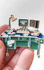 A miniature desk-themed enamel pin held in a person's hand against a light gray background. The pin features a blue desk with green drawers, gold-tone metal outlines, a silver desk lamp, a computer monitor, and a speaker.