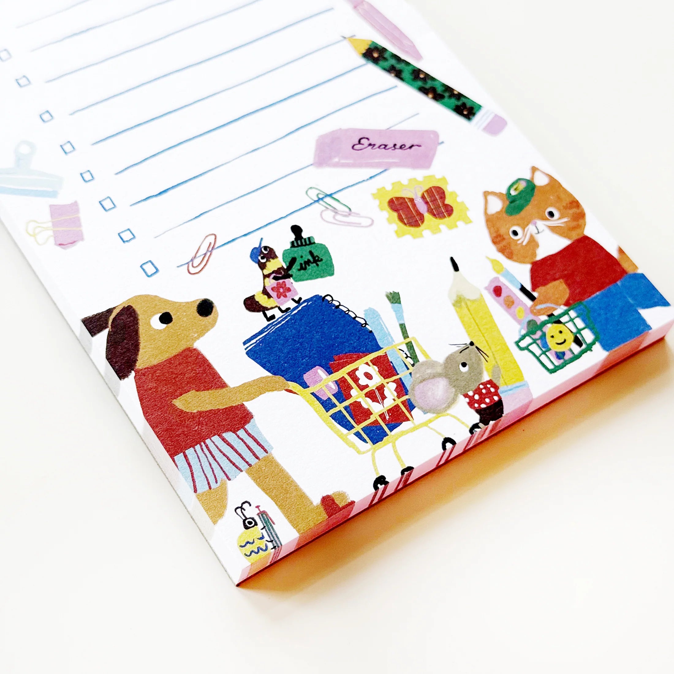 A close-up of a white notepad with a colorful cartoon illustration of a dog pushing a shopping cart and a cat holding an eraser, alongside lined writing areas and a small pencil drawing.