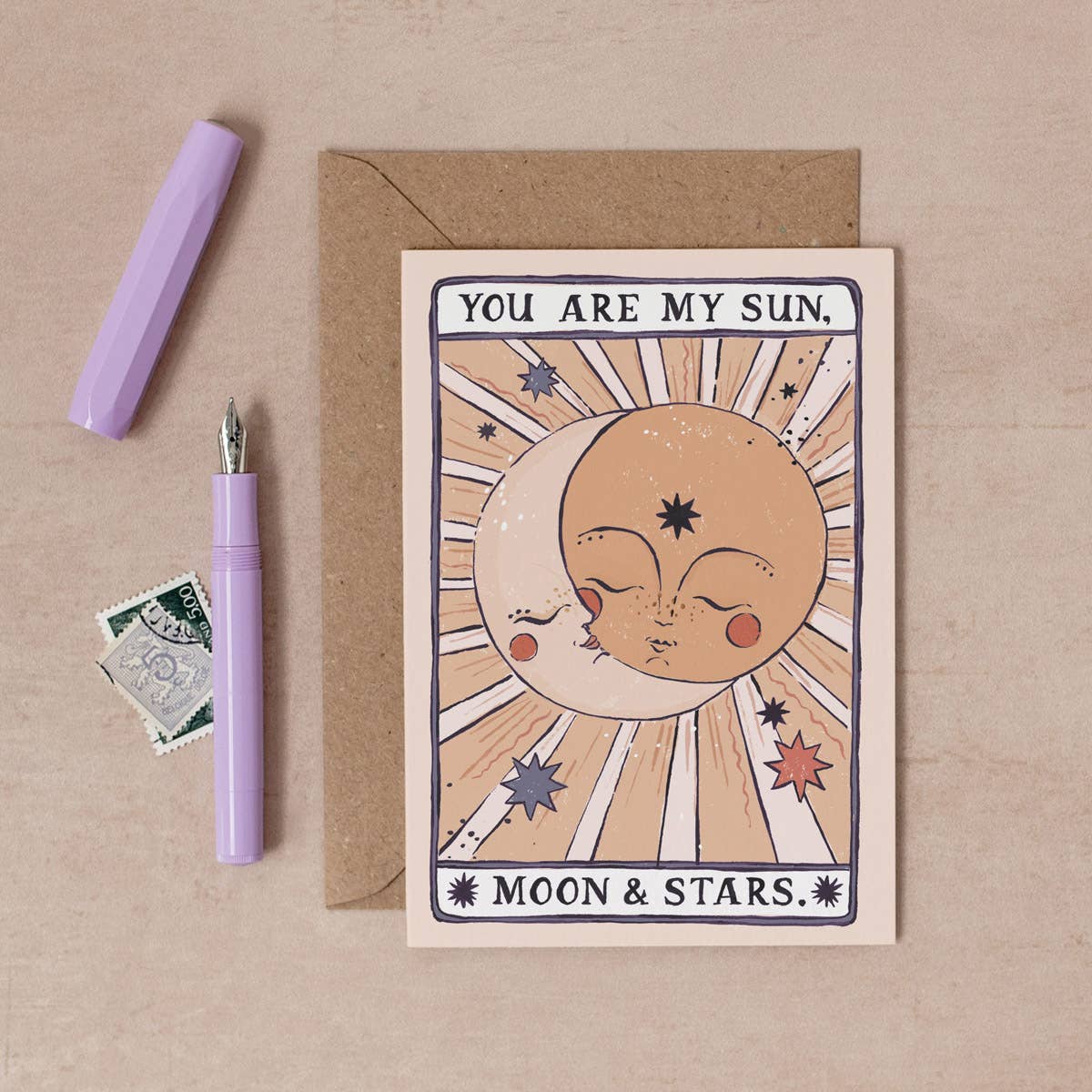 Greeting card with moon and sun design, pen, and envelope on a beige background
