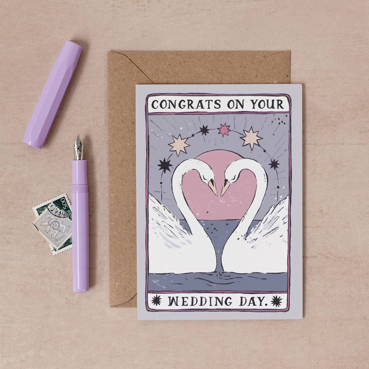 Wedding card with swans and 'Congrats on your wedding day' text, next to a pen and envelope on a beige background