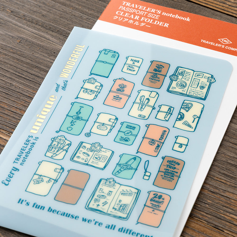 A sheet of decorative stickers featuring illustrated travel backpacks and passport-style holders in pastel blue, peach, cream, and teal, placed on a wooden surface. The sheet includes the text 'TRAVELER'S notebook' and 'PASSPORT SIZE CLEAR FOLDER' on an orange header, with the brand mark 'TRAVELER'S COMPANY' visible.
