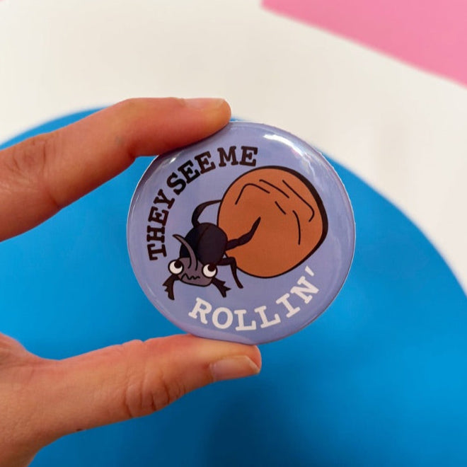 Button badge with text 'THEY SEE ME ROLLIN'' held by a hand against a colorful background