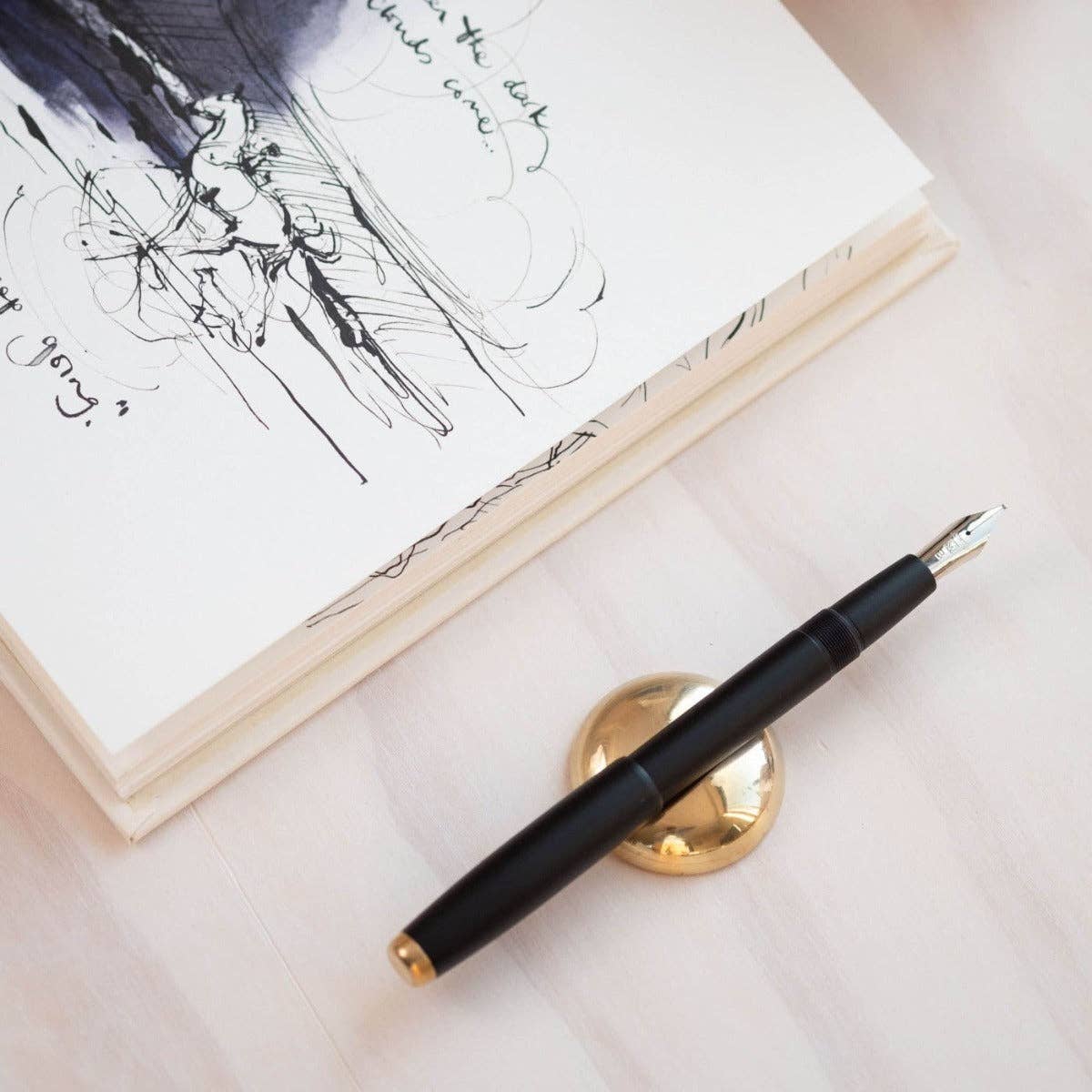 Black fountain pen with gold accents on a white surface next to a book with sketches.
