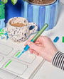Hand holding a green marker over a drawing with a cup of coffee and stationery items in the background.