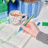 Hand holding a green marker over a drawing with a cup of coffee and stationery items in the background.