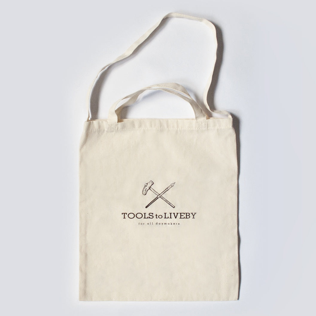 Beige tote bag with 'TOOLS TO LIVE BY' logo on a light gray background