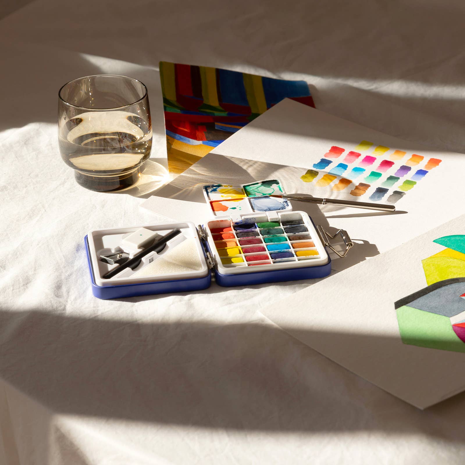 Art supplies including watercolor paints, a glass of water, and a color chart on a white surface.
