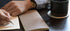 Person writing in a notebook with a black mug on a table